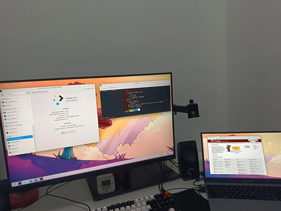 Desk setup with a large monitor and a laptop running FreeBSD with KDE Plasma. The main screen shows KDE system information and a terminal, while the laptop displays the FreeBSD website.