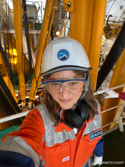Selfie in safety gear — Melanie wearing a JAMSTEC hard hat, safety goggles, and an orange coverall takes a selfie among yellow drilling machinery on the Chikyu.