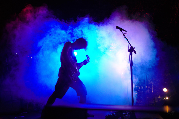 Slightly blurry photo of a bass player silhouetted against a smoke cloud lit by blue and purple lights.