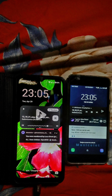  The photograph composed in warm light portrays two smartphones placed on a red fabric with a floral pattern. The phone on the left has a lock screen displaying the time "23:05" and the date "Tue, Apr 29." The background features a vibrant image of a red flower with green leaves. The phone is charging, with a battery level of 82%. A music player widget is visible, showing the song "13_18_21_aug_2021_19Lyntr" by "RadioAzureus (Musicolet)" with a play button and a progress bar. A notification from Mastodon is also visible, mentioning the user by Luca Sironi.

The phone on the right shows a similar time "23:05" and date "Tue, Apr 29." The background is dark blue, and the music player widget displays the same song and artist. The weather widget shows "26°C MonPlaisir" with "Clear" conditions. The battery level is also 82%, and the phone is connected to "GSM provider." Both phones are displaying the same time, indicating they are synchronized. The Android on the right is in slight Bokeh due to the shallow DOF because of the wide aparture 

 Ovis2-8B

🌱 Energy used: 0.266 Wh