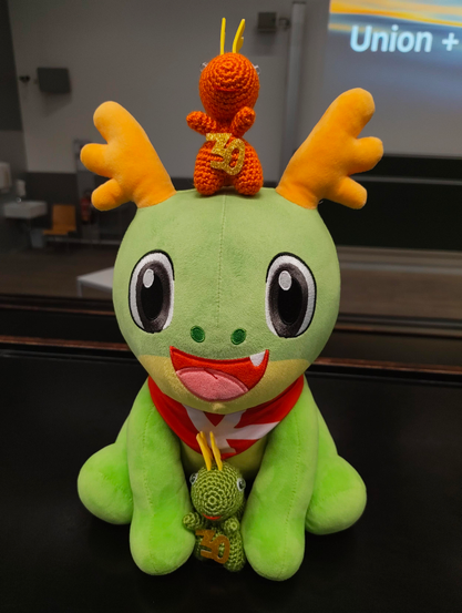Konqi plushie with a green amigurumi KDE dragon between the arms and an orange amigurumi KDE dragon on top of it.